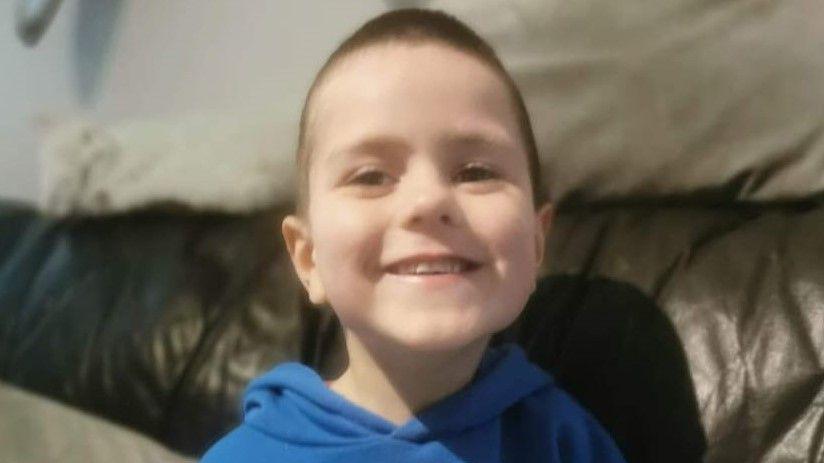 Kyran Durnin - a young boy with short brown hair smiles at the camera while sitting on a dark leather sofa. He is wearing a blue hoodie.