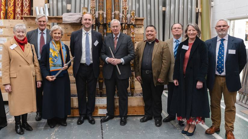 The Duke and Duchess of Gloucester standing with seven people from the Friends of Gloucester Cathedral, in a workshop with organ pipes stacked up vertically behind them