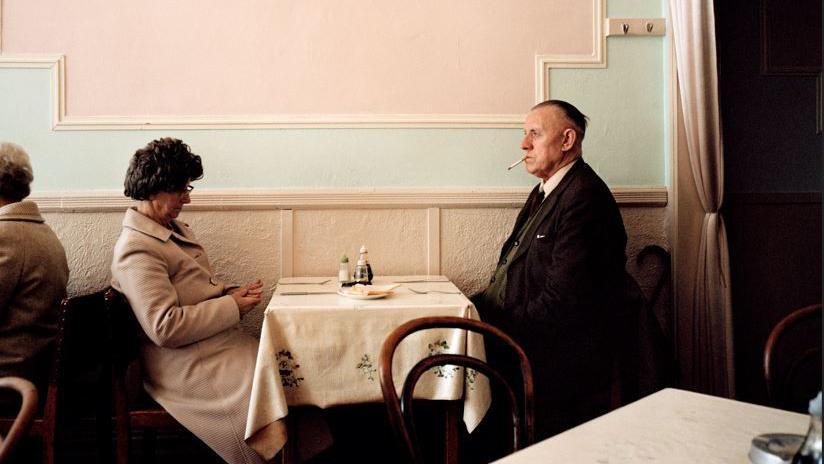 A couple sit in a cafe in the 1960s, one man is smoking a cigarette 