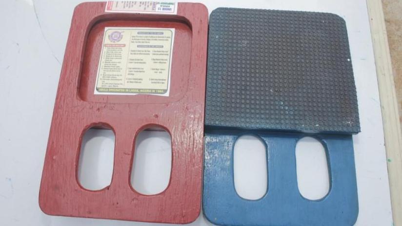 Two rectangular Abula bats are pictured: the red one with a hollowed centre displaying game rules, and the blue one with a textured rubber surface for striking. Both feature twin oval finger grips at the base, a distinctive design unique to the Nigerian court game.