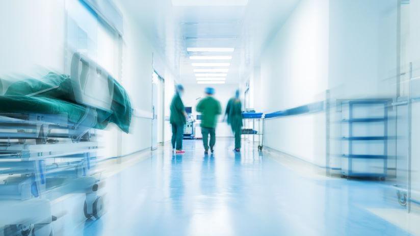 Stock image shows a blurred hospital ward. In the distance you can see the figures of three people wearing scrubs and hats.