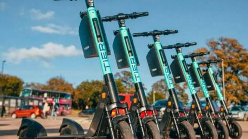 Blue Tier scooters lined up with a red bus and other vehicles in the background.