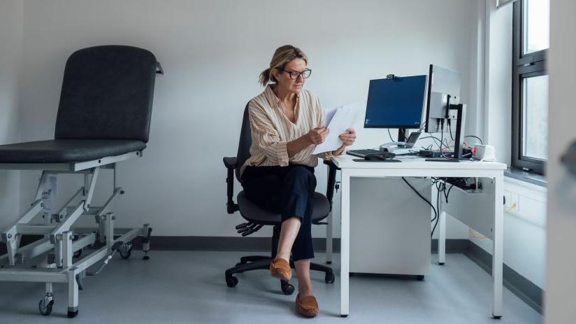 A woman at a computer reads a document on paper. She is sitting in an officer chair and there is a medical bed behind her.