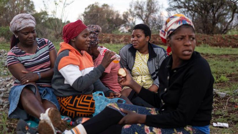 Five women sit close together on the grass as they wait for the illegal miners to emerge. 