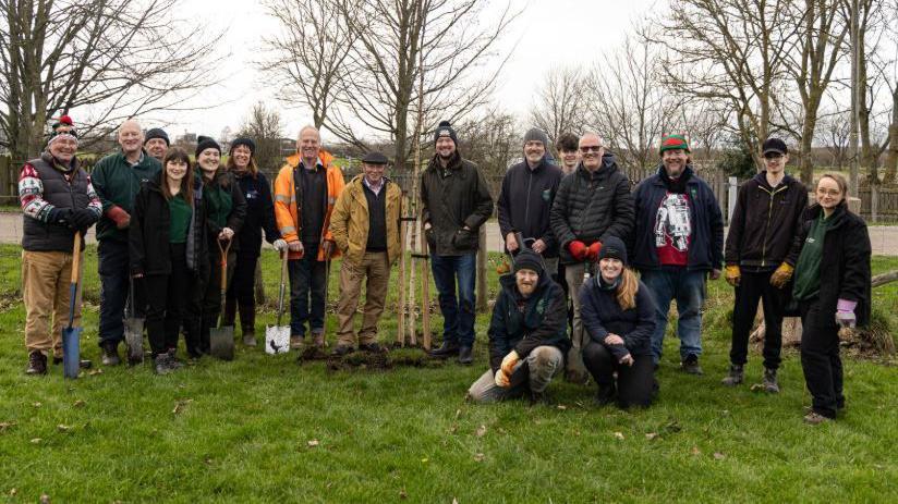 A group of 17 people dressed up warm standing next to a tree that has just been planted. They are standing on grass, several holding spades, with other trees seen in the background.