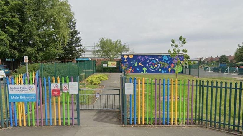 A green, yellow, purple and blue-coloured metal fence bearing the main entrance sign to St John Fisher RC Primary School, with a gate in the centre opening on to a paved walkway towards a school building. A car park can be seen to one side in the distance, with a playground the other side.