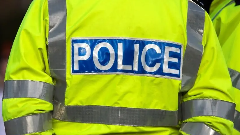 A police officer is standing with their back towards the camera. They are wearing a yellow hi-vis jacket that says Police on the back in white writing. 