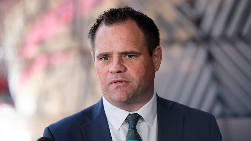 Irish Minister of State Neale Richmond, with short brown hair, wearing a navy suit, white shirt and dark green tie. The background of the image is blurred.