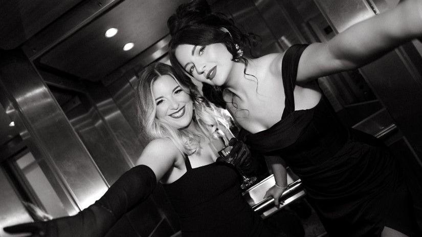 Georgie Mitchell and Beth Smith are pictured here in a lift. The photograph is black and white. Georgie has long blonde hair and wears a black dress. Beth has dark hair which is pinned in a bun. She too is wearing a black dress.