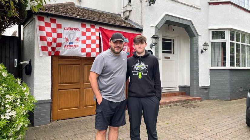 Phil Cross, 40, who has a grey t-shirt, baseball cap and dark blue shorts, stands next to his son who has a black hoodie and jogging bottoms. Behind them are two Liverpool flags hanging on the garage of their semi-detached house.