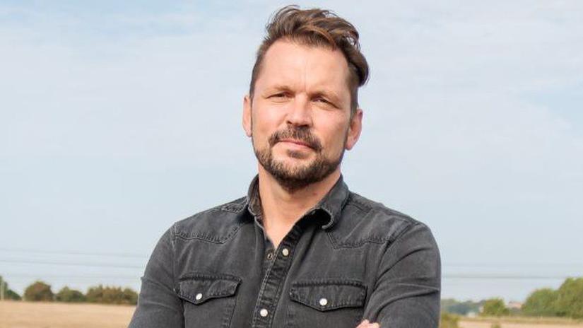 A man stands in a field on a cloudy day. He has his arms folded in front of him. He has brown hair that is swept to the right hand side and he is wearing a faded black denim shirt. He has a brown beard.
