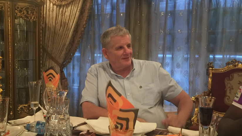 Grey haired man in a pale blue short sleeved shirt smiles to someone to one side while sitting at a table dotted with wine glasses and plates