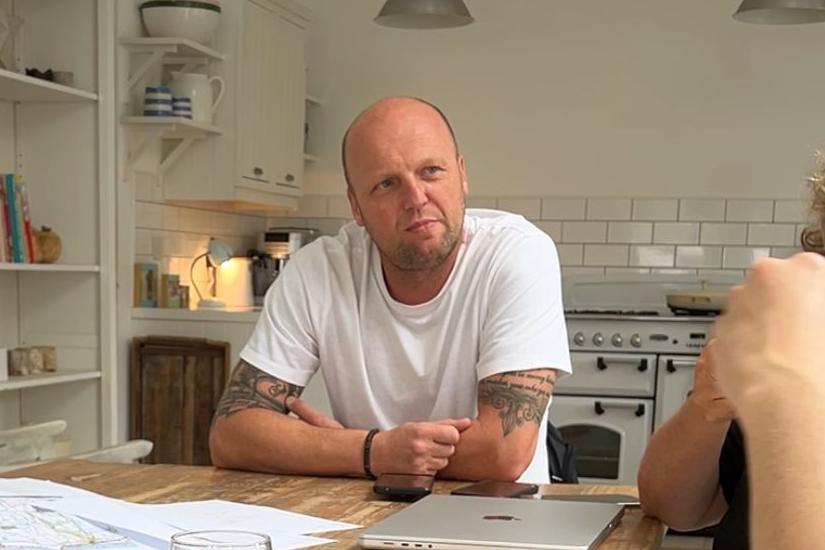 Ian is pictured wearing a white t shirt and sitting at a table. He has a shaved head and tattoos on his arms.