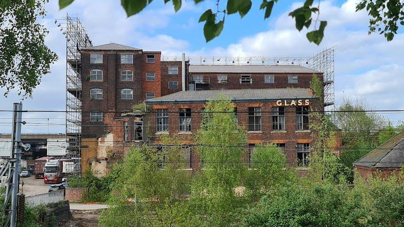 A brown brick building with scaffolding at the side. At the front of the building is a metal fence and some green trees. The word 'Glass' is at the top of building.