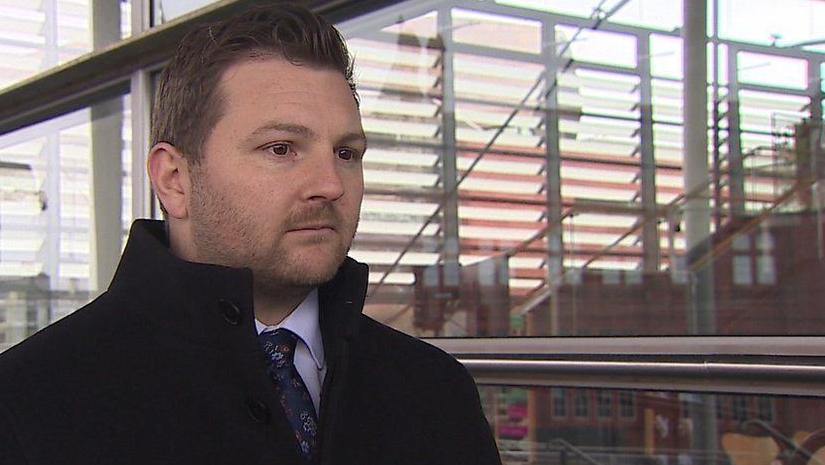 Samuel Kurtz in a dark jacket and tie standing in front of the Senedd foyer