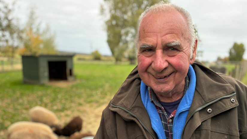 Dave Buscombe wearing a brown jacket, blue fleece and shirt. He is smiling and in the background is a green field and some blurry pigs.