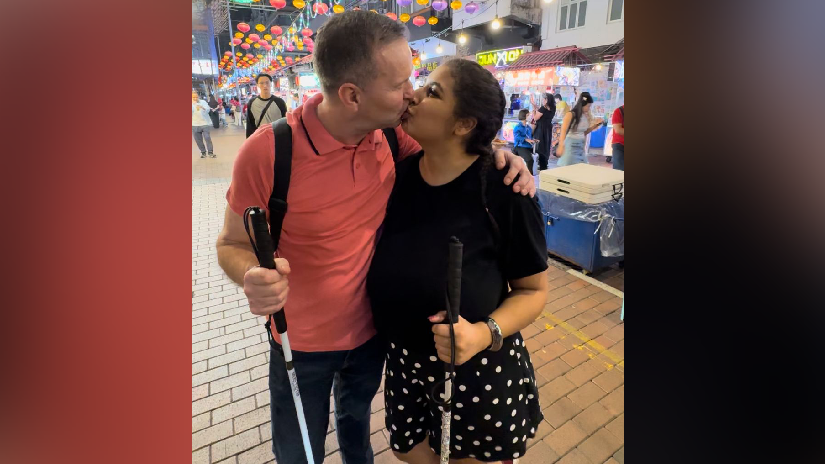 Nicola and Paul woman kissing on a street in east Asia. They are both holding canes as they have visual impairments. He is wearing blue jeans and a salmon pink shirt, and she is wearing a black top and a black and white polka dot skirt.