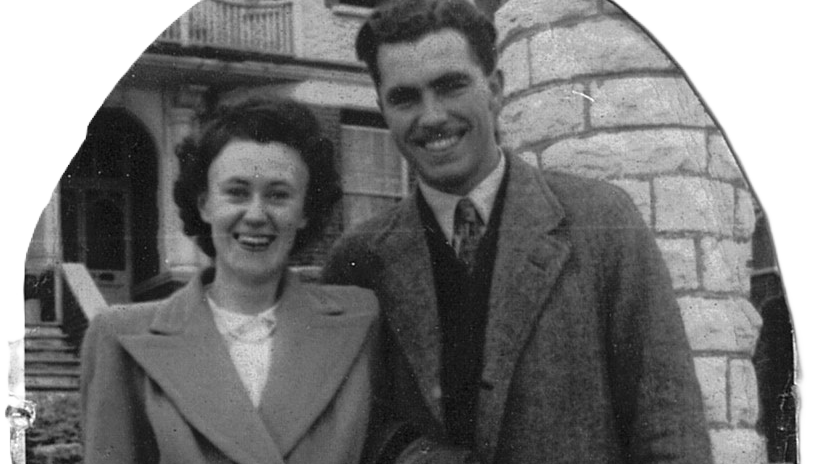 A black and white cutout family photo of Pam and Charles, they are standing outside a brick building and both smiling at the camera. Pam is on the left, she has short dark hair fashioned into a blow dried perm, a padded shoulder coat with wide lapel collar and white blouse with a single string of pearls as a necklace. Charles is on the left, he has short dark hair and a moustache, wears a collared coact with a shirt and tie and v neck jumper.