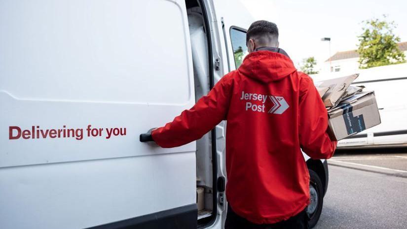 A man with a red Jersey Post jacket with his hand on a white van that says 'Delivering for you'.