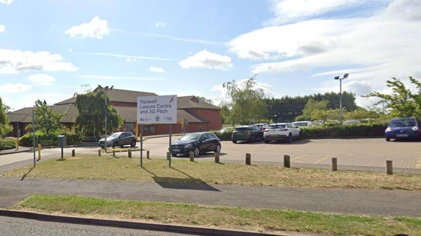 Entrance to leisure centre with "Redwell Leisure Centre and 3G Pitch" sign in the foreground.  There is a car park behind the sign and a two-storey brick building to the left, flanked by trees