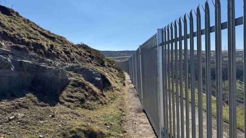 A long, spiky metal fence on the right and a rocky mountain on the left, with the blue sky above.