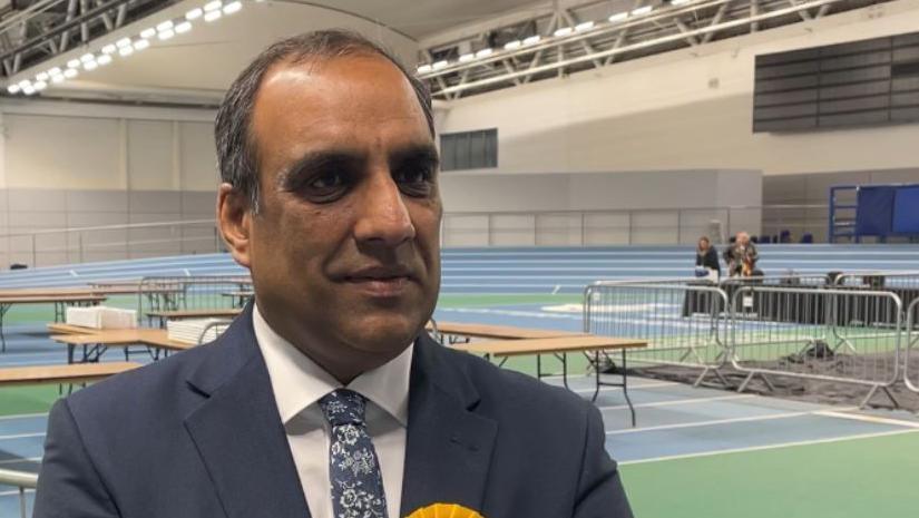 Mr Mohammed, an Asian male, stands in a sports hall wearing a blue suit, white shirt and floral tie