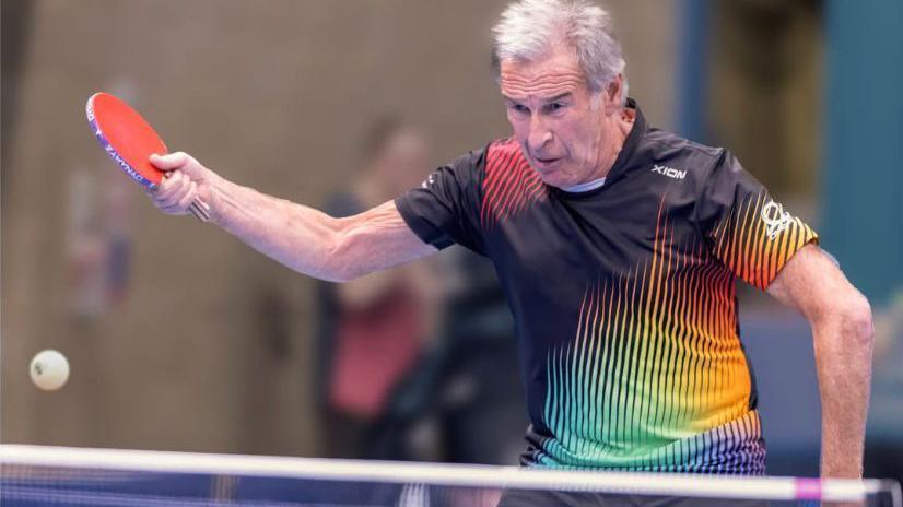 A table tennis player mid-swing in a multi-coloured top, his face is a picture of concentration and the ball is on its way across the table.