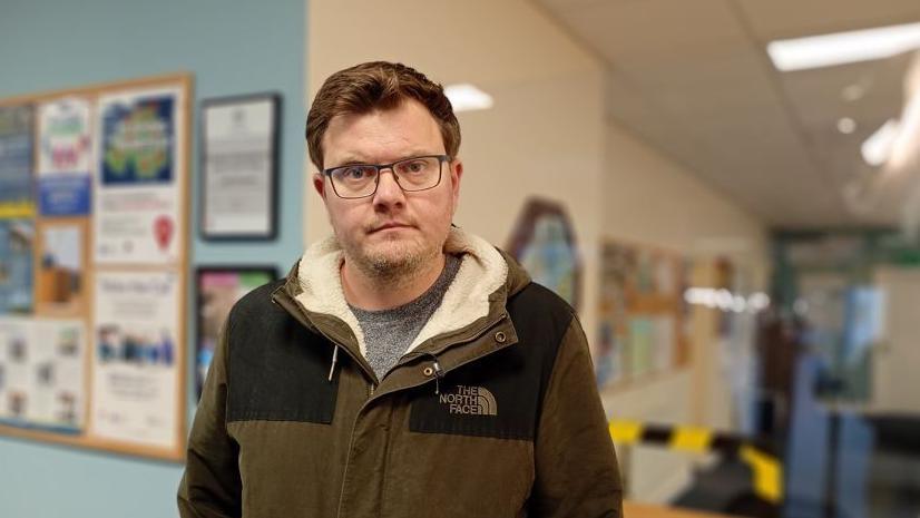 Aaron Skinner, a man with brown hair and wearing a brown and black jacket, with grey top. He has glasses on his face.