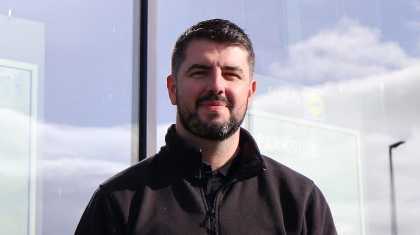 Declan has short black hair and facial hair, a beard and moustache. He is standing in front of a shop window, as he wears a black zip-up fleece and top.
