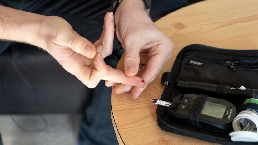 A man conducts a finger prick test to check his insulin levels. On the right of the image is a black case containing digital equipment and a tube of swabs. On the left of the image is the mans finger with a spot of blood.