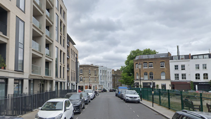 A street view of Faraday Road, with a modern block of flats on the left and a park and play area on the rights, with cafes on the corners of a crossroads visible in the distance