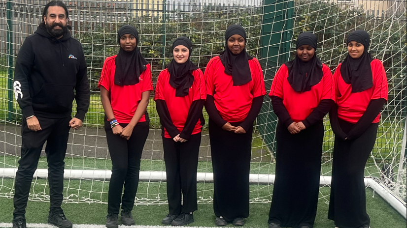 Five school pupils in sports hijabs stand next to their male teacher inside a football net