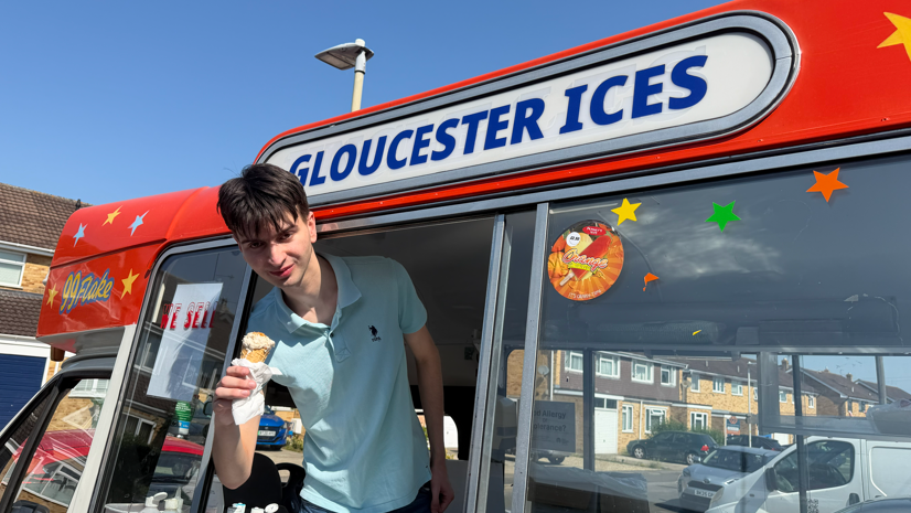 Emery, a young man in a blue polo neck shirt, smiles while holding an ice cream out of the window of an ice cream van, beneath a sign reading: 'Gloucester Ices'