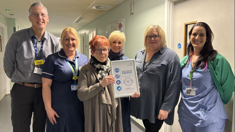 A woman holds a framed certificate, which says "award" as a man and four women stand behind her. They are in the corridor of a hospital. The man stands taller than the others, wearing a grey shirt with a blue lanyard. The women mainly have short hair, ranging in shades from blonde to red to black. Some of them wear hospital scrubs.