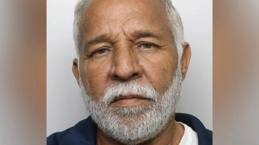 Piran Ditta Khan frowning with a light grey beard and light grey hair. He is stood in front of a light grey background and wearing what appears to be a white t-shirt and blue jacket. 