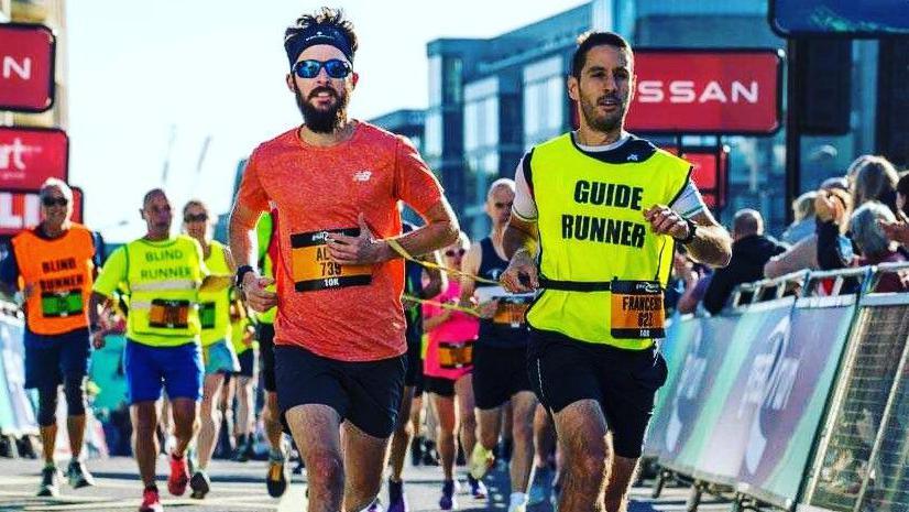 Alex Iness taking part in a running race, running alongside a male guide. Alex is to the left and is wearing glasses, a headband, a red T-shirt and black shorts. The guide next to him has a high-vis top on, is wearing black shorts and a white and black top. There are other runners behind them and spectators to the right, standing behind a metal barrier. 