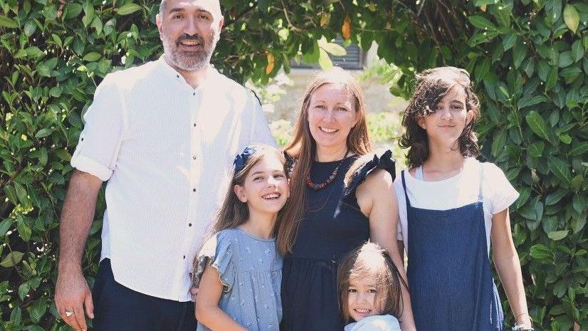 A family of five smiles for a photo taken outside, with a lush green bushy archway, in a garden. A man, a woman and their three children. One child looks teenage-aged, the others are younger.