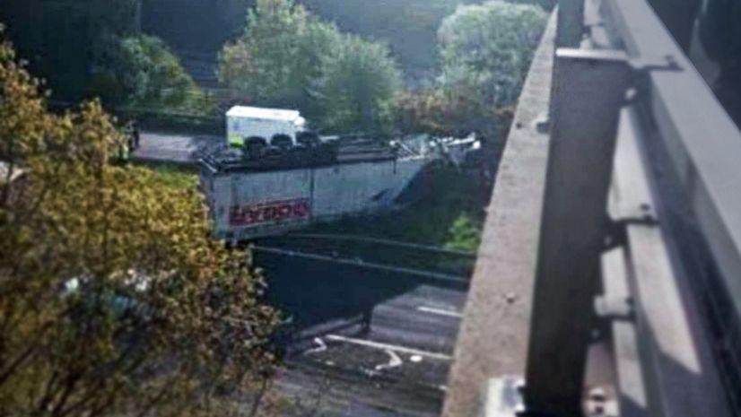 A white HGV trailer lies overturned on a grass embankment beside some trees, view looking down from above