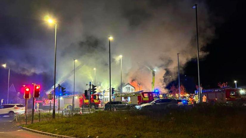 A corner of a junction with traffic lights on red to the left, three fire engines can be seen with water hoses shooting up in the night-time sky, which is thick with smoke and fire from a pub in the background