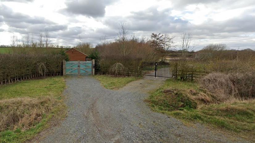 A path leading up to two gates, one is wooden and one is black railing. Both gates lead to fields, with a small wooden structure in the background.