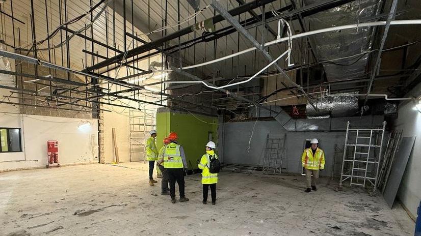 A number of people stand in a large room that is undergoing construction work. They are all wearing hi-vis clothing and hard hats. 