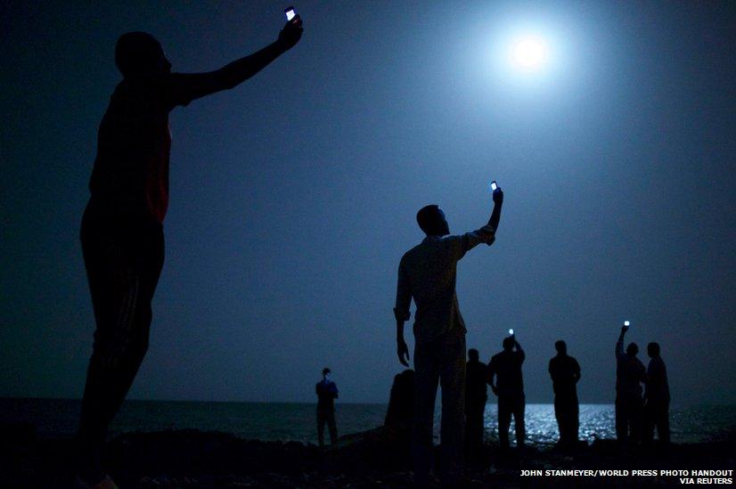 African migrants on the shore of Djibouti city at night, raising their phones in an attempt to capture an inexpensive signal from neighbouring Somalia