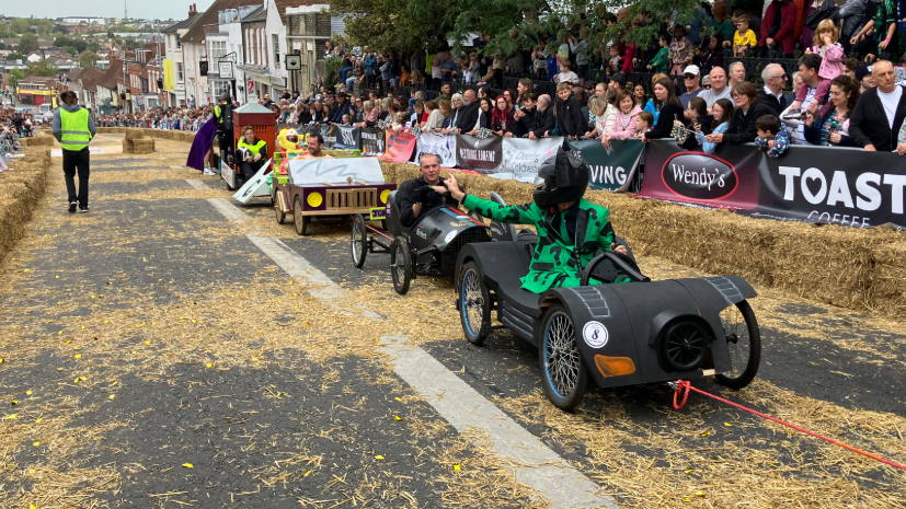 Different coloured karts are being dragged up the hill by a red piece of rope. People are sat in the karts which are on a road. People are also standing behind barriers watching. 