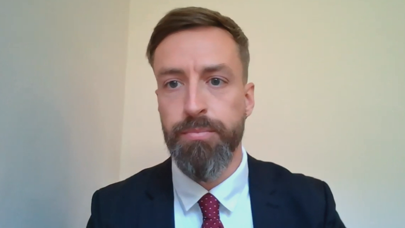 Liam is looking at the camera through Zoom with a plain white background. He has well-kept short brown hair in a quiff, and a full beard that is greying near his chin. He is wearing a black suit, white shirt and red spotted tie.
