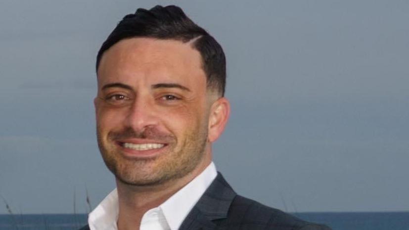 A headshot of Kyle Plesa smiling at the camera. He has black hair and facial stubble.