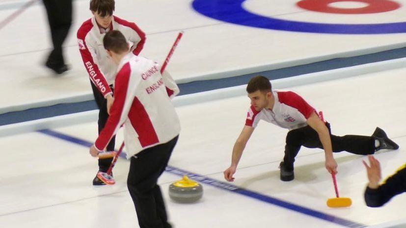 Callum McLain, in England white top with red short sleeves, lets go of the grey granite curling stone, with a yellow handle on top, as he slides down the ice with teammates either side ready to sweep.