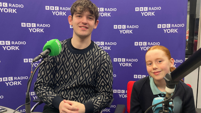 A 20-year-old man and 13-year-old girl sit in front of microphones in a radio studio, smiling at the camera.