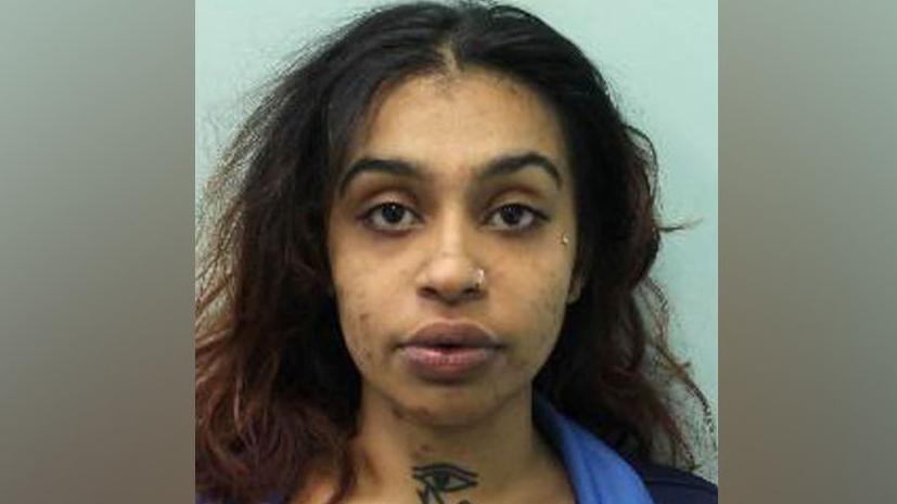 A woman with a neck tattoo and long dark hair looks at the camera for a police custody shot.
