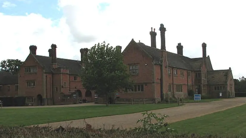 An exterior of an old manor house. The brick building, in extensive grounds, has two wings, with several chimney stacks on each of them.