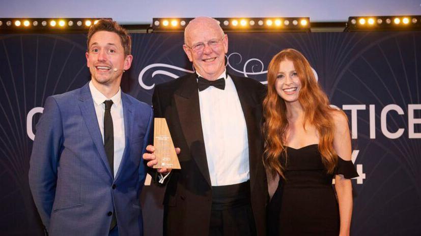 An elderly, bald man in a black tuxedo and glasses holding a wooden award and smiling at the camera with a younger man in a navy suit and a younger, ginger woman standing next to him.